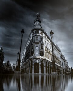 Black and white photograph of the Four Seasons building in Madrid under dramatic grey clouds, with its reflection on the ground — taken by George Adamides, author